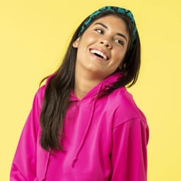 A woman smiles while wearing a vibrant pink hoodie, complemented by a green headband, against a yellow background.