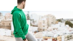 A green hoodie worn by a man standing against a backdrop of a city and ocean. The hoodie features a logo.
