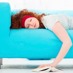 A woman with curly red hair rests on a turquoise couch, her head on the armrest, with a book lying on the floor.