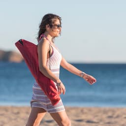 A woman wearing sunglasses walks along the beach, carrying a red chair with a striped dress and the ocean in the background.