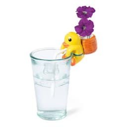 A yellow duck-shaped drink bottle with a small flower pot on its back, holding purple flowers, beside a glass of water.