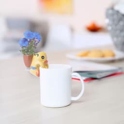A white mug with a yellow duck-shaped planter holding blue flowers. The scene includes a plate of cookies in the background.