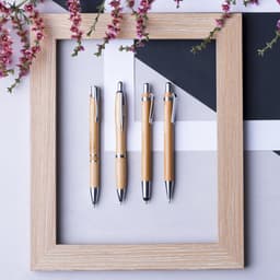 Four wooden pens with silver accents are displayed inside a light wooden frame, surrounded by pink flowers.