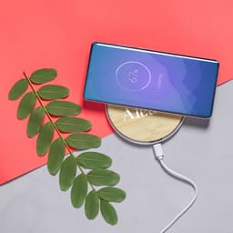 A wireless charger with a wooden base and a smartphone displaying charging status, accompanied by a green leaf on a grey surface.
