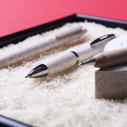 A collection of pens in grey and cream, resting on white rice in a black tray. The display includes a branded pen.
