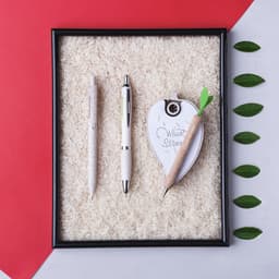 A framed display featuring three pens: a wooden pencil, a silver pen, and a carrot-shaped pen, arranged on a rice background.