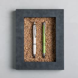 A black-framed display featuring two pens: one white and one green, placed on a bed of grains.