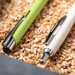 A green and a white pen are resting on a bed of oat grains, showcasing their sleek designs. Each pen features a logo.