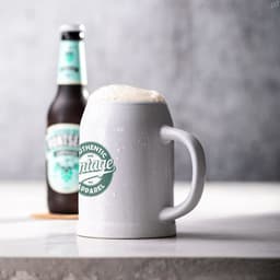 A white ceramic beer mug with a handle, filled with frothy beer, beside a beer bottle. The mug features a logo.