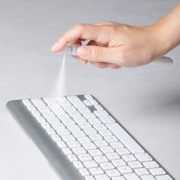 A hand spraying a cleaning solution onto a silver and white keyboard. The spray creates a fine mist.