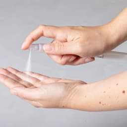 A hand holding a spray bottle, applying a fine mist onto an open palm. The bottle is clear and has a nozzle.