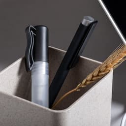 A container holding a black pen and a clear marker, alongside some decorative wheat, set against a neutral background.