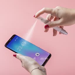 A hand holding a white pen sprays a fine mist over a smartphone screen against a pink background.