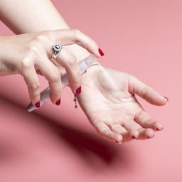 A hand holding a spray pen with a clear body, spraying onto an open palm against a pink background.