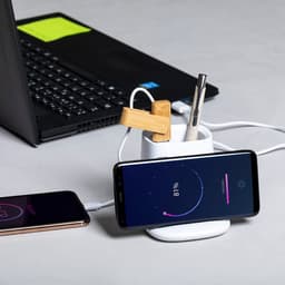 A white multifunction pencil holder with wooden clips, a pen, and a smartphone charging on a wireless charger beside a laptop.