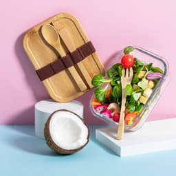 A bamboo lunch box with utensils sits beside a glass container filled with a fresh salad and a coconut on display.