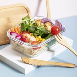 A glass container filled with a salad featuring shrimp, tomatoes, corn, and various vegetables, with wooden cutlery beside it.