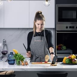 A woman in a grey apron prepares food with a knife and cutting board on a kitchen countertop, surrounded by fresh ingredients.