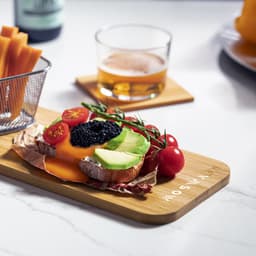 A bamboo chopping board features avocado, tomatoes, and caviar on top of bread, accompanied by a drink and snacks.