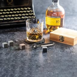 A crystal glass sits on a coaster with grey stone ice cubes, a bottle of whisky, and a wooden box nearby.