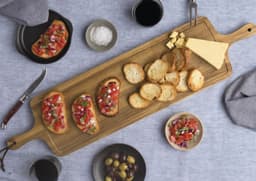 A wooden tapas serving board features bruschetta, crackers, cheese, olives, and drinks on a textured blue tablecloth.