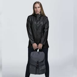 A person wearing a black jacket holds a grey backpack with a flap closure and a logo.