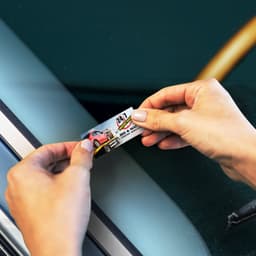 A hand applying a colourful sticker to a car window, featuring a graphic of a car and a logo.