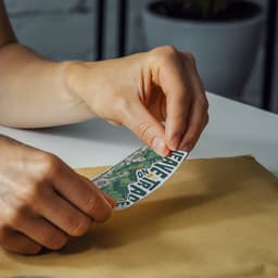 A hand is peeling a colourful sticker from a backing, with a textured brown surface underneath.
