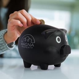 A black piggy bank with a logo featuring "MAGNA PROPERTY" is shown as a hand inserts a coin into it.