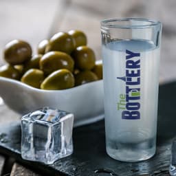 A shot glass featuring the logo "The BOTTLERY," accompanied by a bowl of olives and a cube of ice on a slate surface.