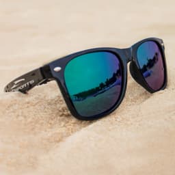 Black sunglasses with mirrored green and purple lenses, resting on sand. The sunglasses have a branding logo on the side.