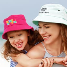 A pink bucket hat and a light blue bucket hat worn by a mother and child, both smiling at the beach.