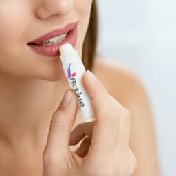A lip balm tube in white and colourful branding, held near a person's lips, with a smooth, creamy balm visible.