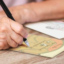 A note pad in soft pastel colours with a hand holding a pen, writing on it. The pad features a design with a logo.