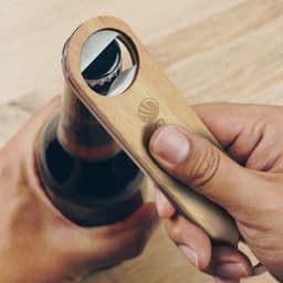 A drink bottle opener made of wood and metal, being used to open a dark beverage bottle. It features a logo.