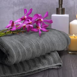A grey bath towel folded neatly with a bouquet of purple orchids, a white container, and a candle in the background.