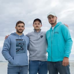 Three men stand together wearing hoodies in various shades of blue and grey. One hoodie features a logo.