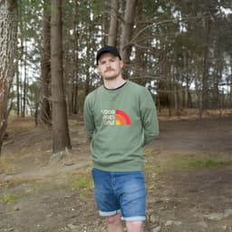 A man stands in a forest wearing a green unisex sweatshirt with a logo. He pairs it with denim shorts.