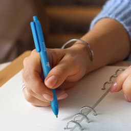 A blue PLA pen being held above a notepad with a hand adorned with rings. The pen has a logo.