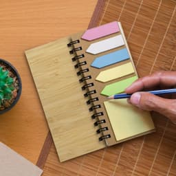A bamboo sticky note wallet featuring sticky note flags in pink, blue, yellow, and green, alongside a pen and a small plant.