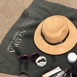 A green waffle towel laid on sand, accompanied by a straw hat, sunglasses, a watch, and a cream jar.