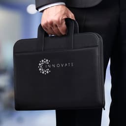 A black travel portfolio case with two handles, featuring a logo on the front, held by a person in a suit.