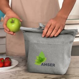 A grey cooler bag with a logo, held by a person, alongside a plate of strawberries and an apple.