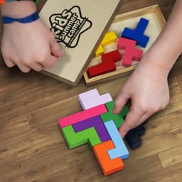 A colourful wooden pentomino puzzle set displayed on a wooden surface, with hands assembling the pieces.