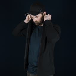 A man in a black Swiss Peak Urban Hoodie adjusts the hood, wearing a black cap. The background is dark.