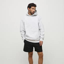 A man wearing a grey hoodie and black shorts stands against a neutral background. The hoodie features a logo.