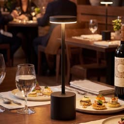 A black rechargeable table lamp stands on a dining table, illuminating an elegant setting with food and drinks.