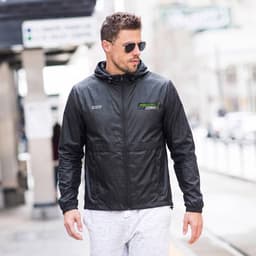 A man wearing a black windbreaker with a hood, paired with sunglasses and light-coloured trousers. The jacket has a logo.
