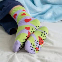 A pair of green and lilac socks with heart patterns and grip dots on the soles, resting on a bed.
