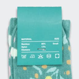A pair of teal kids’ socks featuring a floral design with coloured dots, attached to a material and size label.